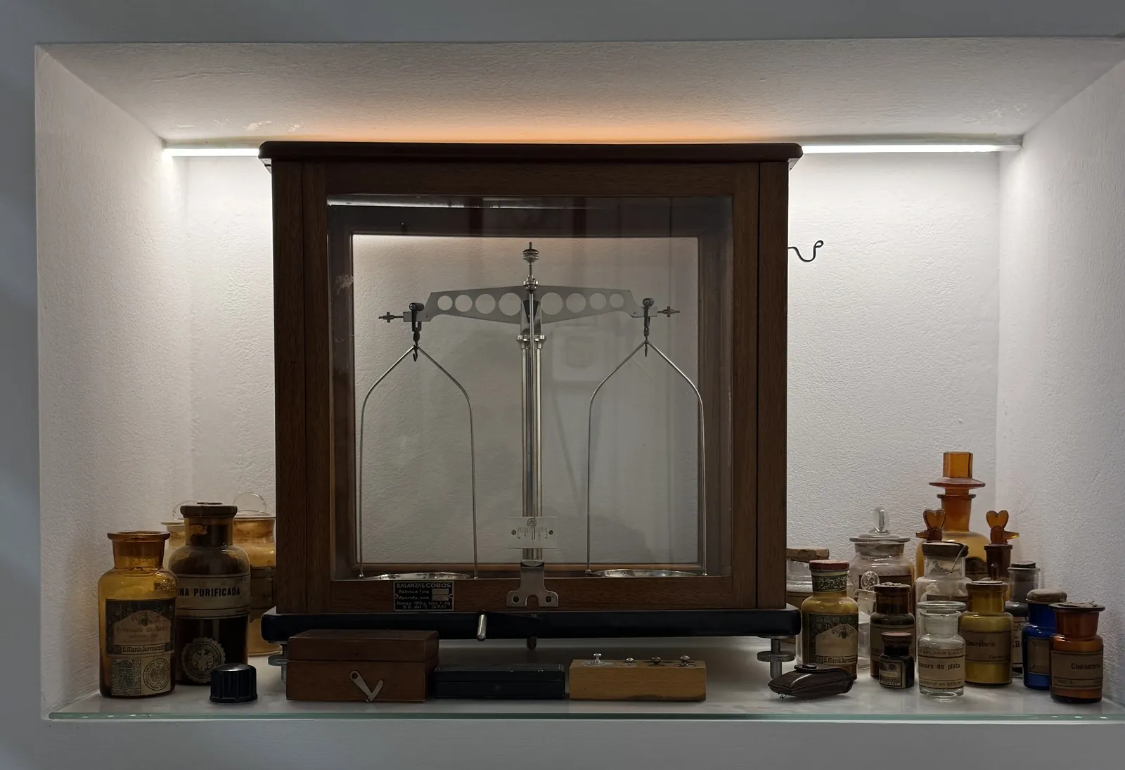 Balanza analítica de platillos en su vitrina de madera, rodeada de frascos de farmacia antiguos